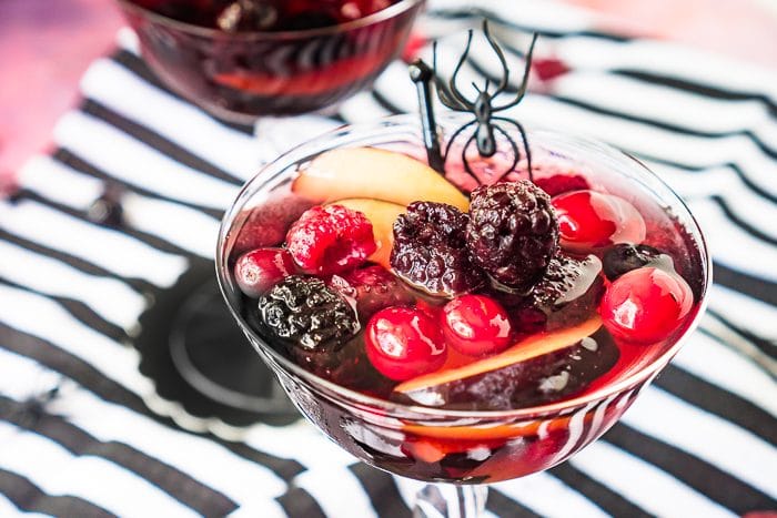A champagne coupe glass filled with a dark sangria and lots of mixed berries, including cranberries. The glass is garnished with a black stir stick and fake black spider. 