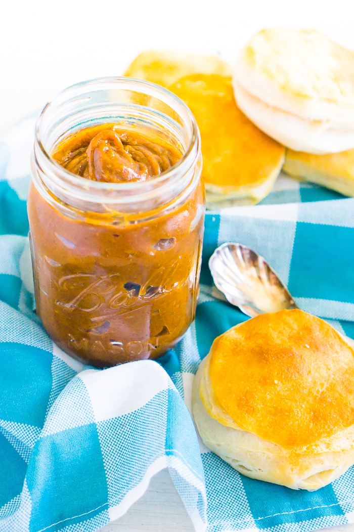 A mason jar full of brown and orange pumpkin butter rest on top of a blue plaid napkin with a pile of biscuits behind it as a small silver spoon laying next to it.