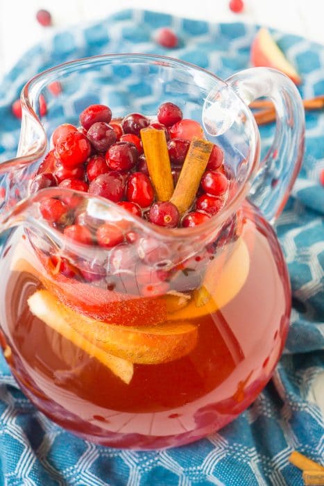 A pitcher filled with a red Thanksgiving punch and topped with apples, cranberries, and cinnamon sticks that is sitting on a teal with white hexagons napkin. 