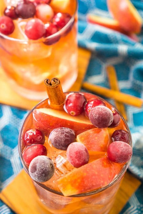 Two short round cocktail glasses sit on wood coasters and are filled with thanksgiving punch and topped with fresh cranberries, apples and a cinnamon stick as a drink stirrer. 