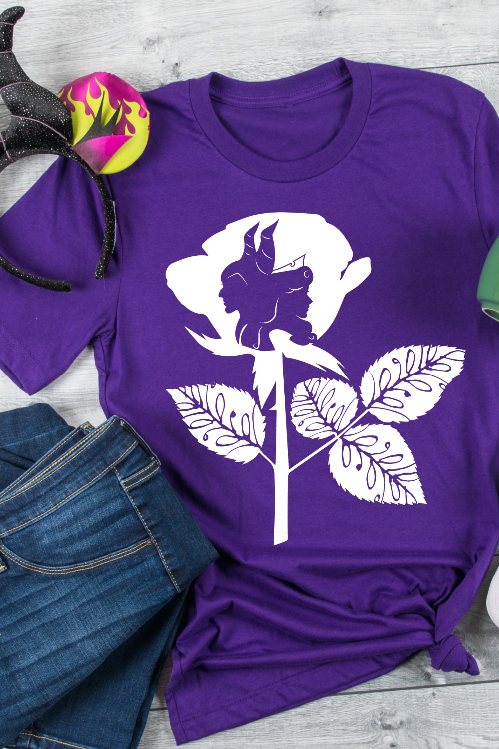 Straight down shot onto a purple tshirt with a white iron on vinyl design on it with Maleficent and Aurora silhouettes standing back to back inside a rose. 