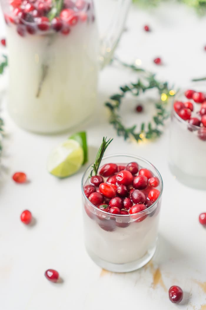 greenery and christmas lights sit on a white wood table with a pitcher of white cranberry margaritas that is also poured into two old fashioned glasses